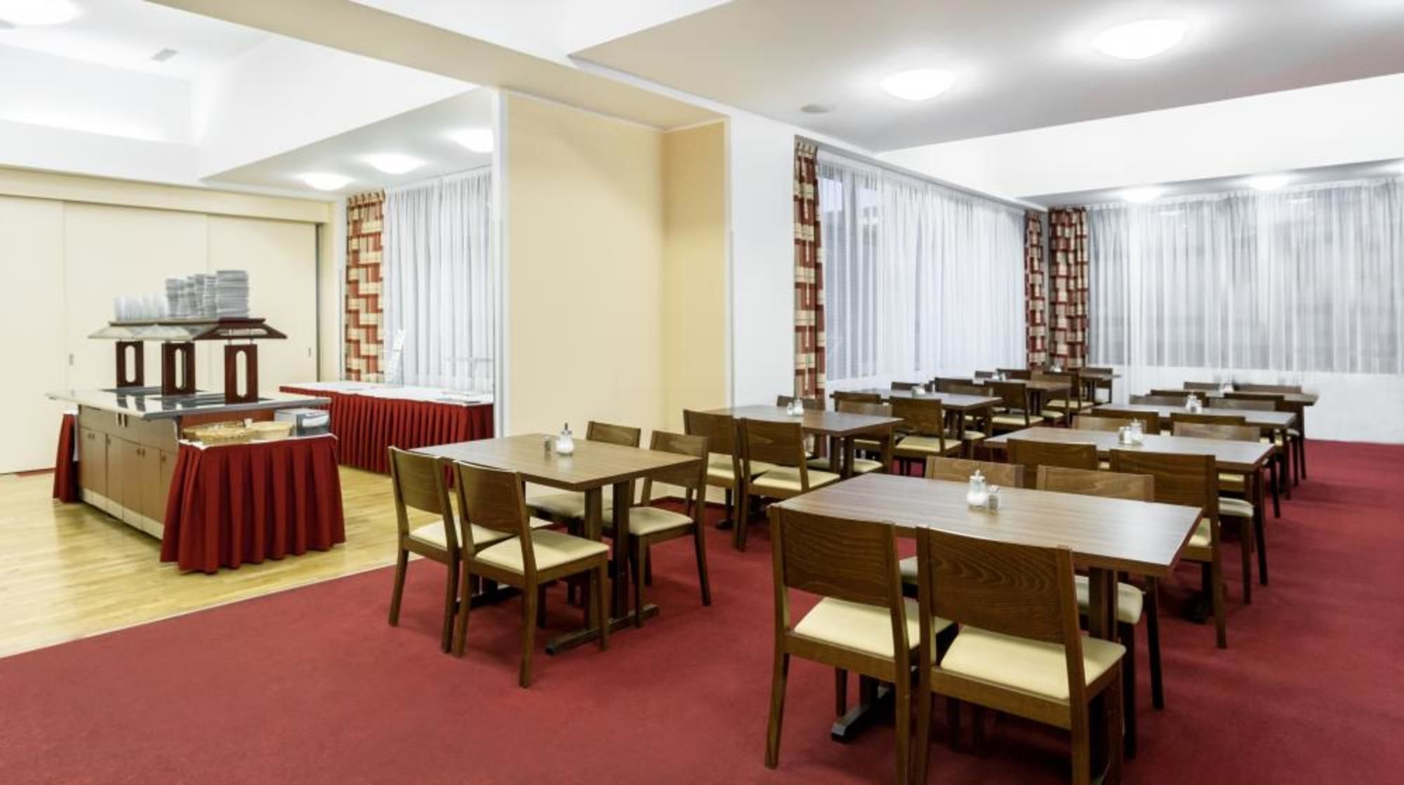 Un salón de conferencias con varias mesas y sillas, una zona de buffet y amplias ventanas con cortinas.