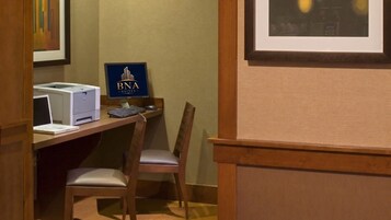A hotel room with a desk, a laptop, a printer, and a chair. There is a framed picture on the wall and a sign with 'BNA Suites' on it.