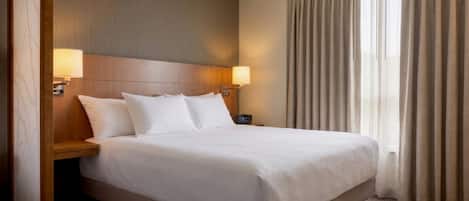 Een hotelkamer met een groot bed, twee bedlampen, een houten hoofdeinde en een raam met gordijnen.