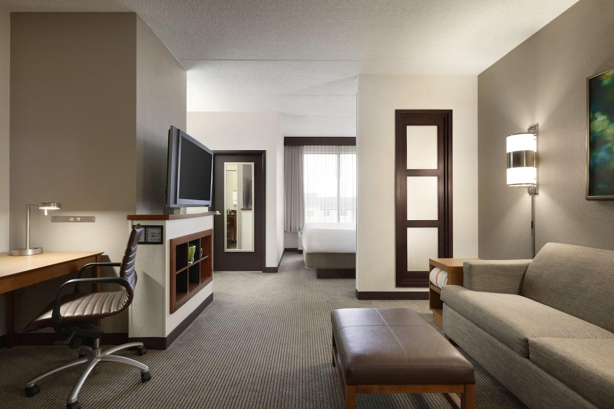 Quarto de hotel com escrivaninha, cadeira, TV, sofá e luminária de cabeceira.