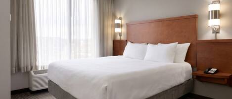 A hotel room with a large bed, a bedside table with a phone, a wall-mounted light, and a window with sheer curtains.