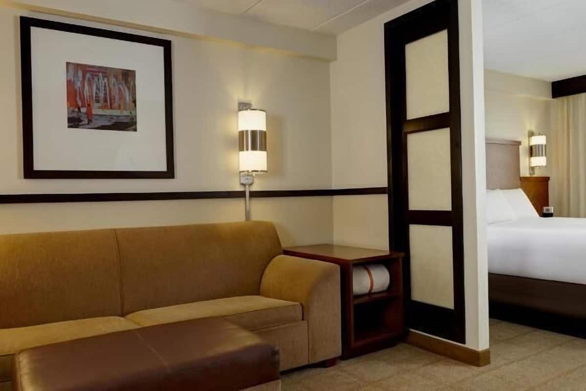 Quarto de hotel com cama, sofá, mesa pequena, abajur e quadro na parede.