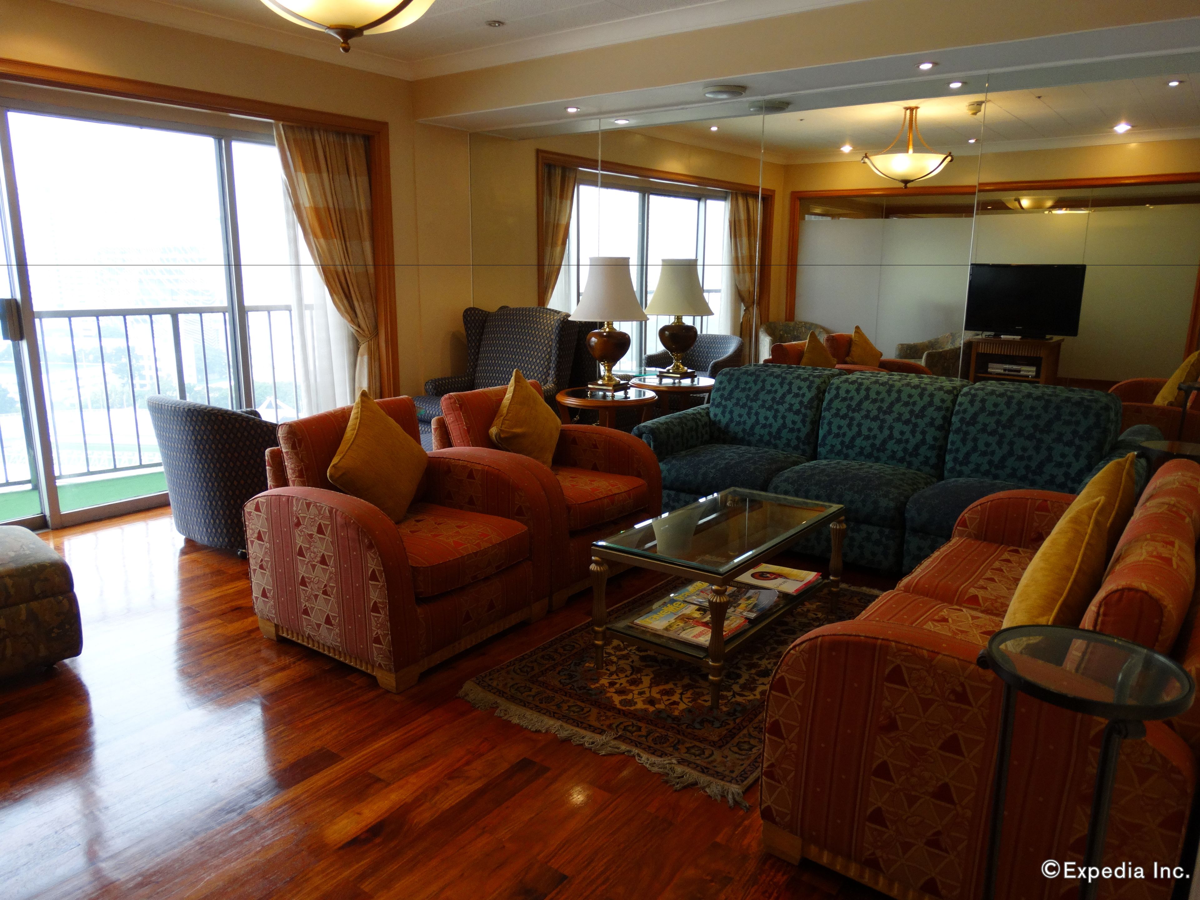 A spacious living room with a large sofa, armchairs, and a coffee table. There is a television and a lamp on a side table.