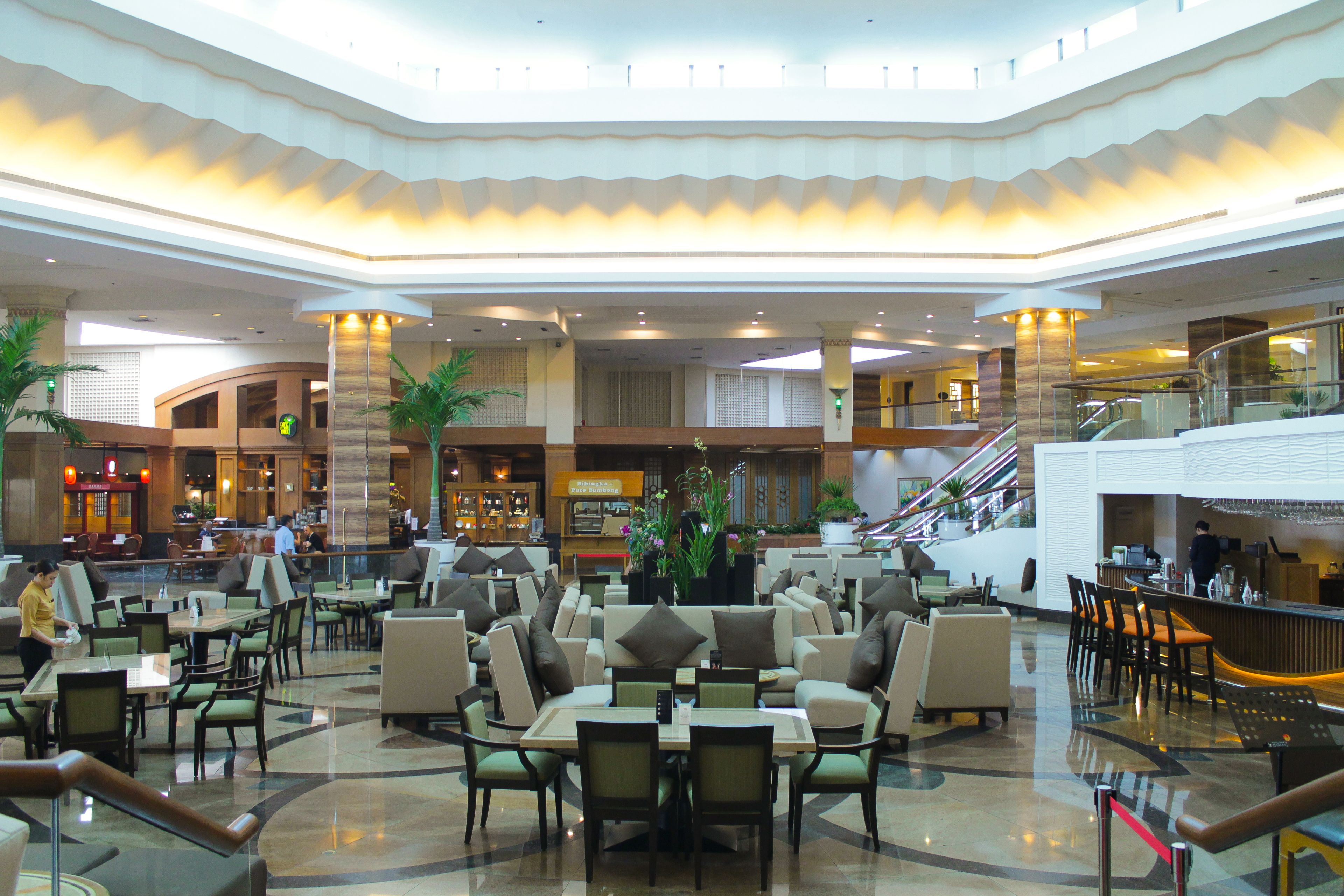 A spacious hotel lobby with multiple seating areas, a central staircase, and a reception desk.