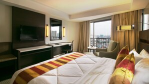 Habitación de hotel con una cama grande, un escritorio, una silla, un televisor y vistas a la ciudad.