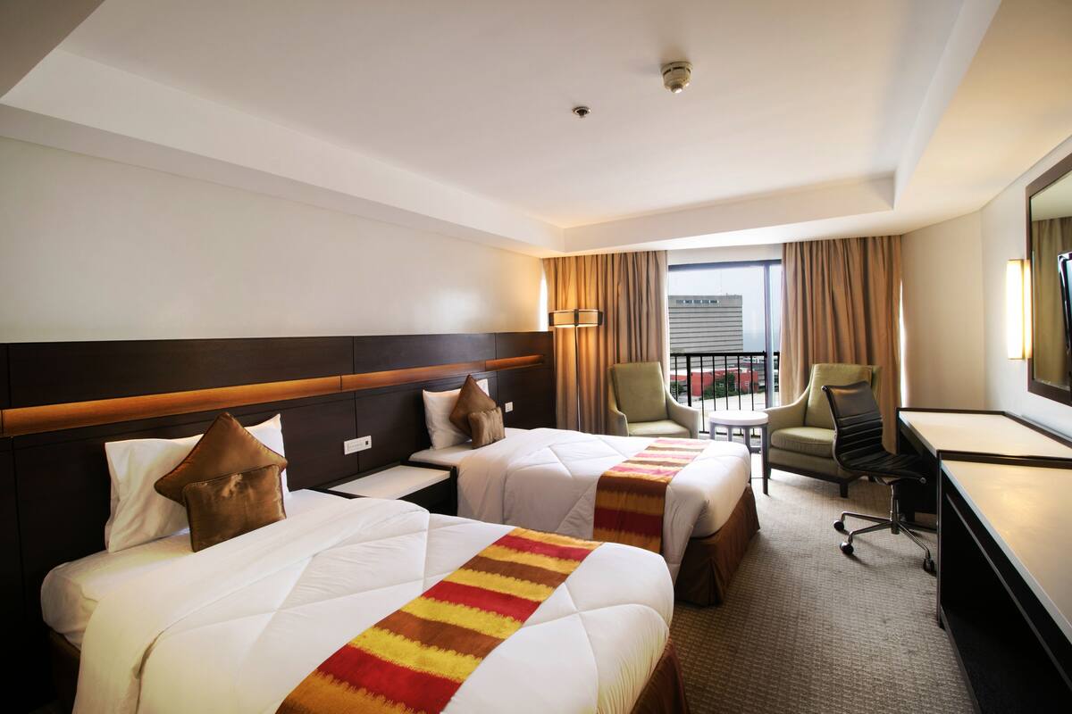 A hotel room with two beds, a desk, a chair, and a view of the outdoors.