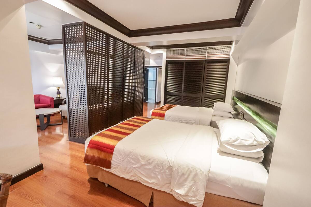 A hotel room with two beds, a wooden floor, a partition wall, and a view of the outside.