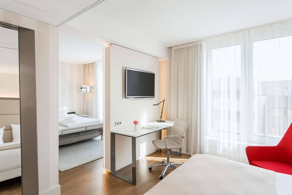 A hotel room with a bed, a desk with a computer, a chair, a TV, and a window with curtains.