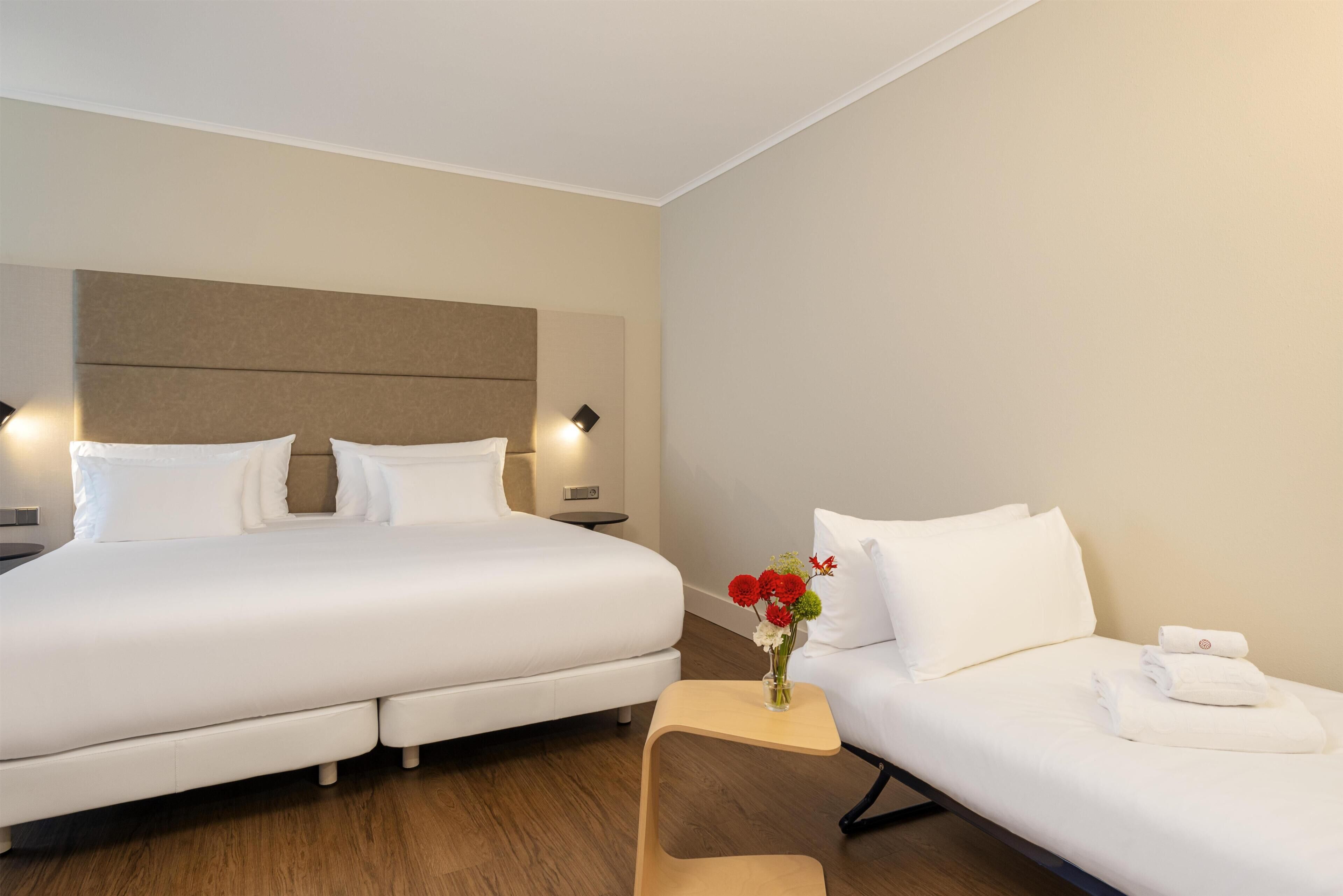 Ein Hotelzimmer mit einem großen Bett, einem kleinen Sofa, einem Nachttisch mit Lampe und einer Vase mit Blumen.