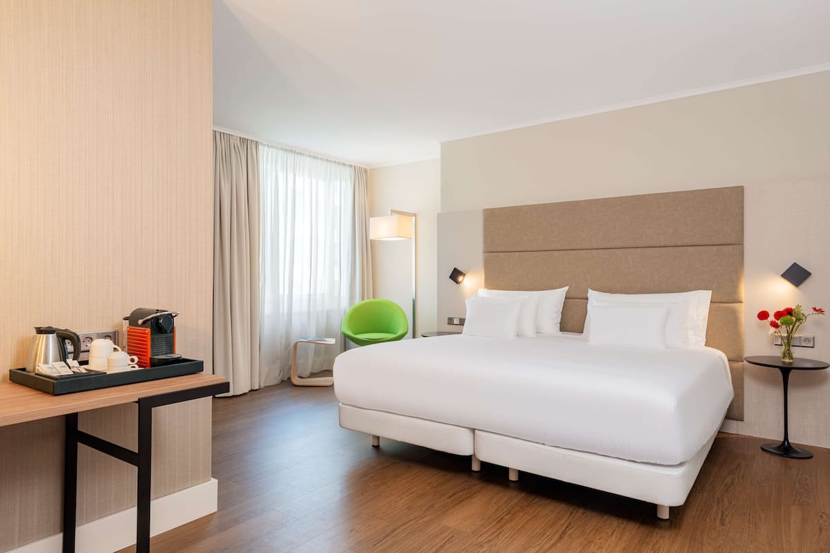 A modern hotel room with a large bed, a desk with a coffee maker, a green chair, and a small table with flowers.