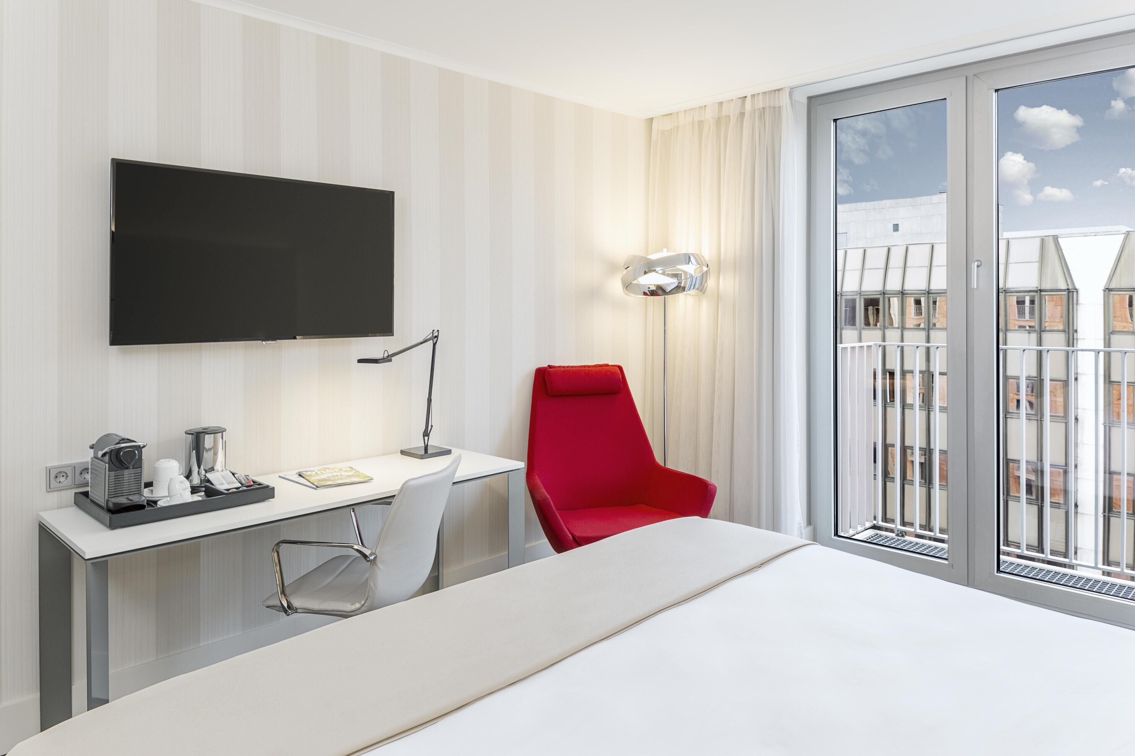 A hotel room with a bed, a desk with a flat-screen TV, a red chair, a desk lamp, and a window with a view of buildings.