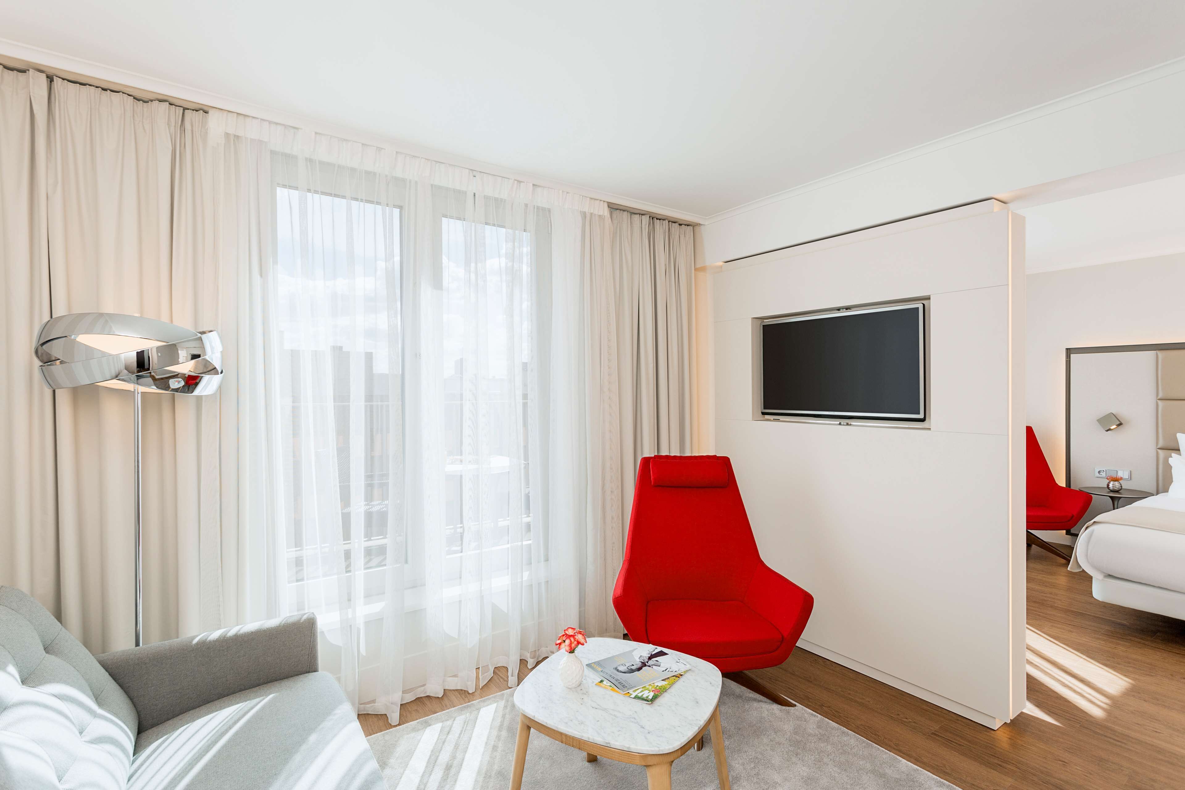 Moderni hotellihuone, jossa on suuri ikkuna, punainen nojatuoli, pieni pöytä, jossa on lehti, ja valkoisilla lakanoilla varustettu sänky.