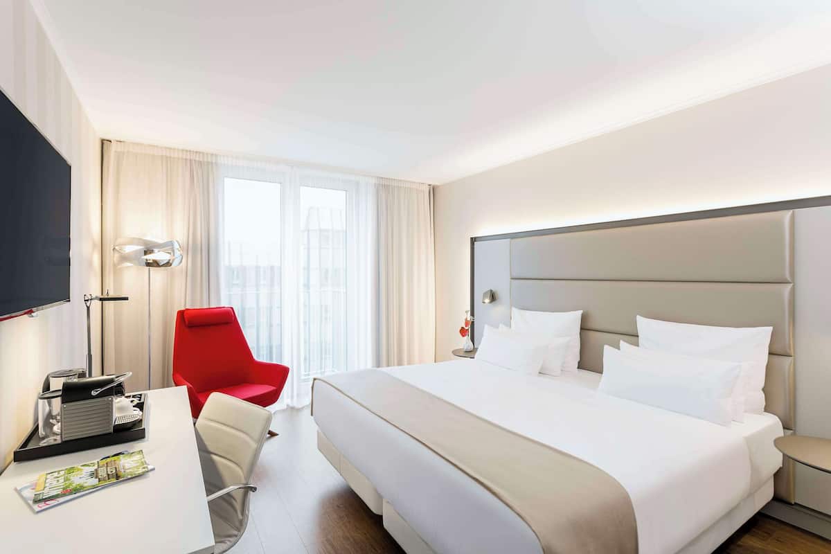 A hotel room with a large bed, a desk with a chair, a red chair, and a TV on the wall.
