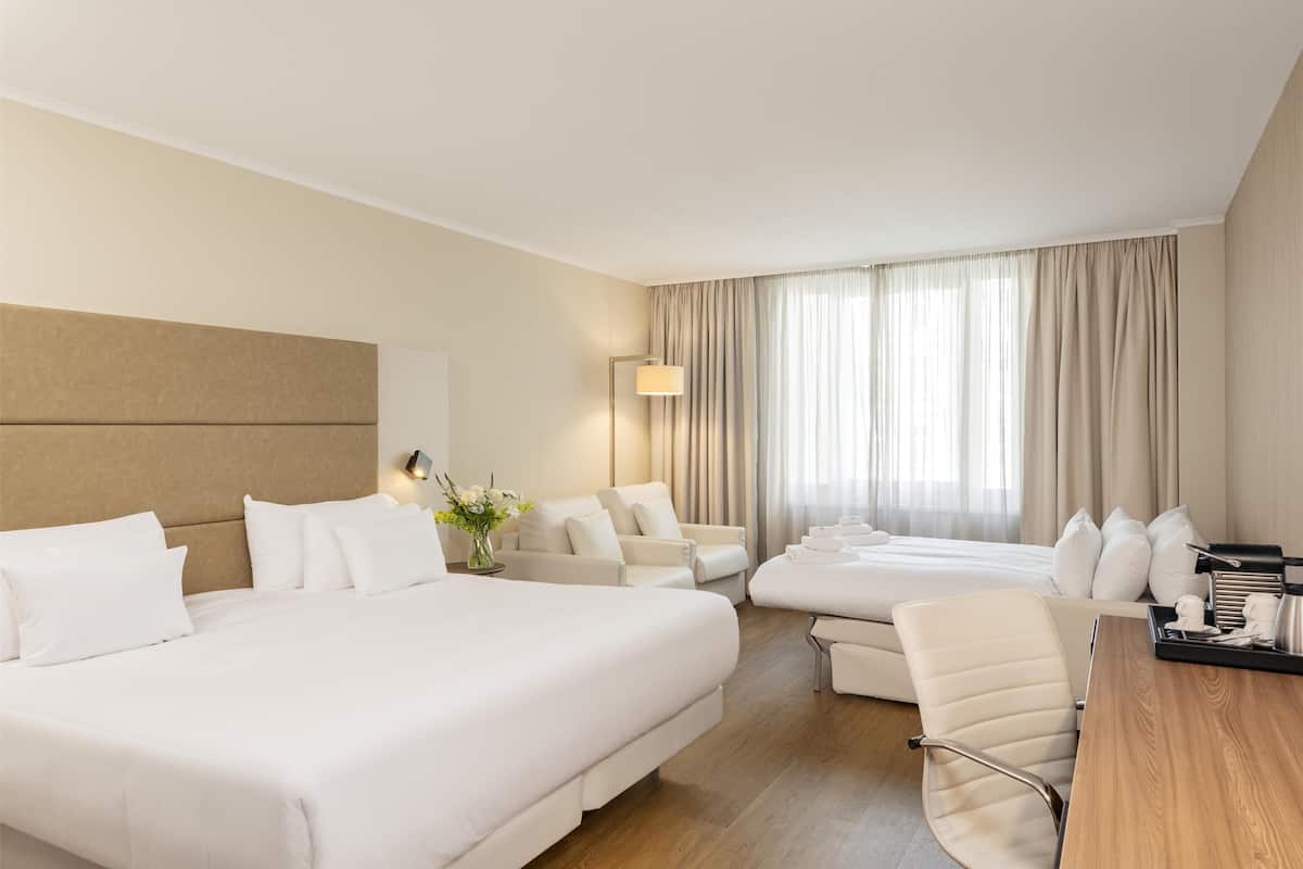 A hotel room with a large bed, a smaller bed, a sofa, a desk, and a lamp.