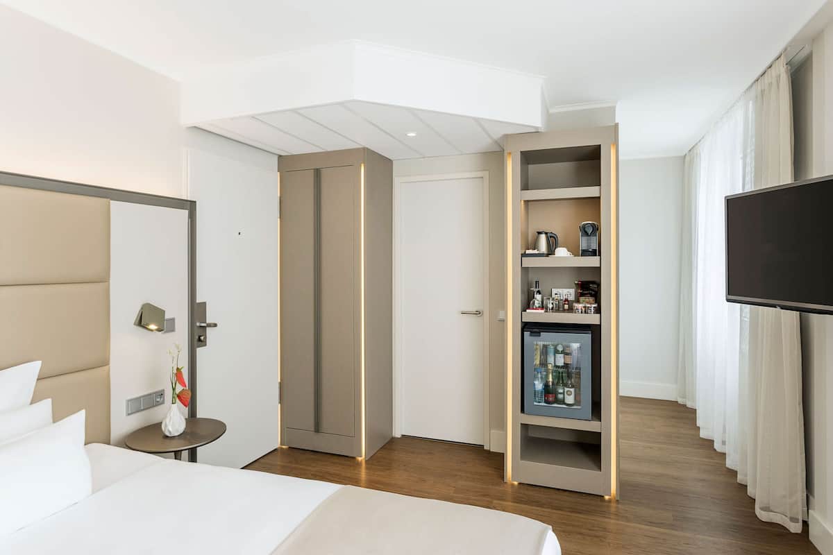 Ein Hotelzimmer mit Bett, Nachttisch, einem kleinen Tisch mit einer Vase, einer Minibar, einem Fernseher und einem Kleiderschrank.