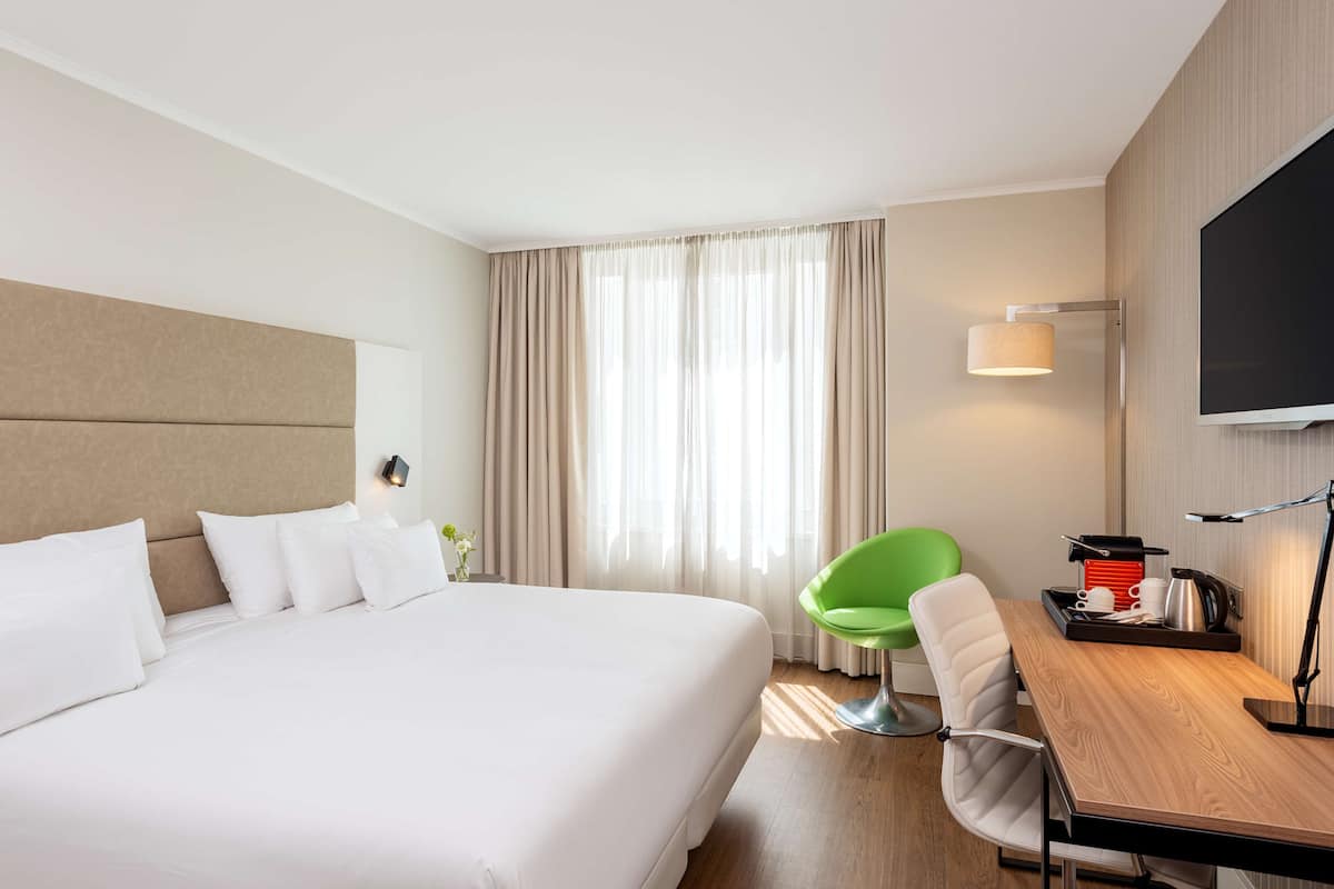 A hotel room with a large bed, a desk with a lamp, a green chair, and a TV on the wall.