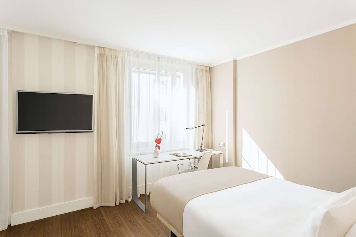 A hotel room with a bed, a desk with a chair, a flat-screen TV, and a window with curtains.
