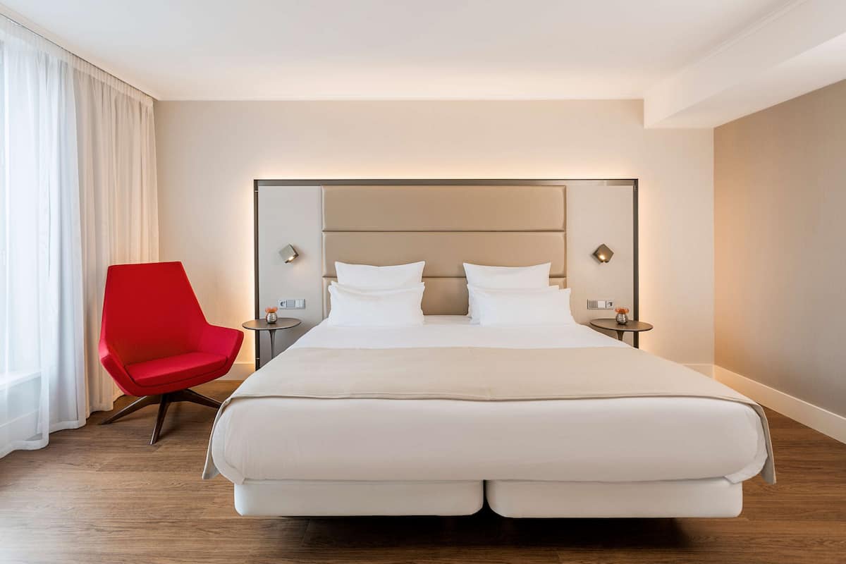 A modern hotel room with a large bed, a red chair, and a nightstand with a lamp.