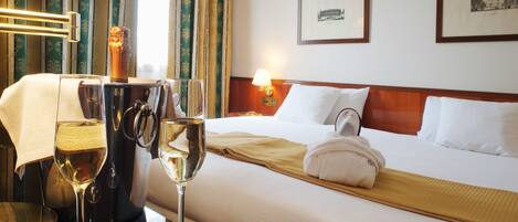 Quarto de hotel com cama bem arrumada, mesa de cabeceira com abajur, taça de vinho e uma garrafa.