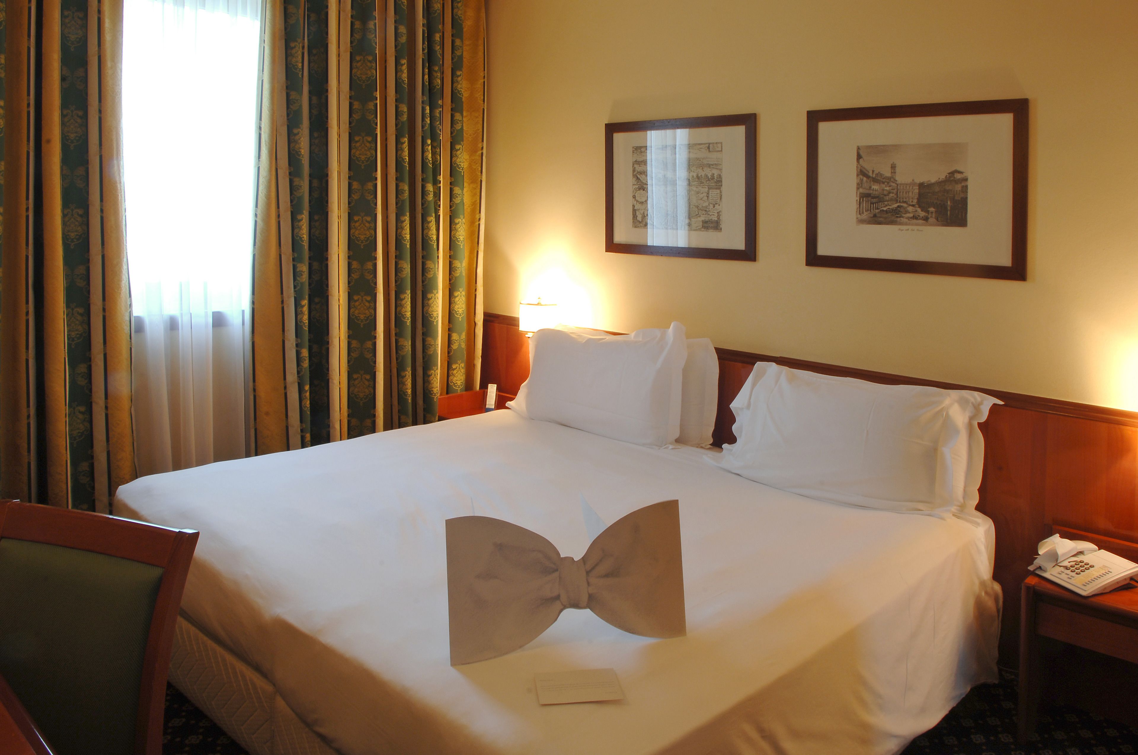 A hotel room with a neatly made bed, a chair, a bedside table with a lamp, and two framed pictures on the wall.
