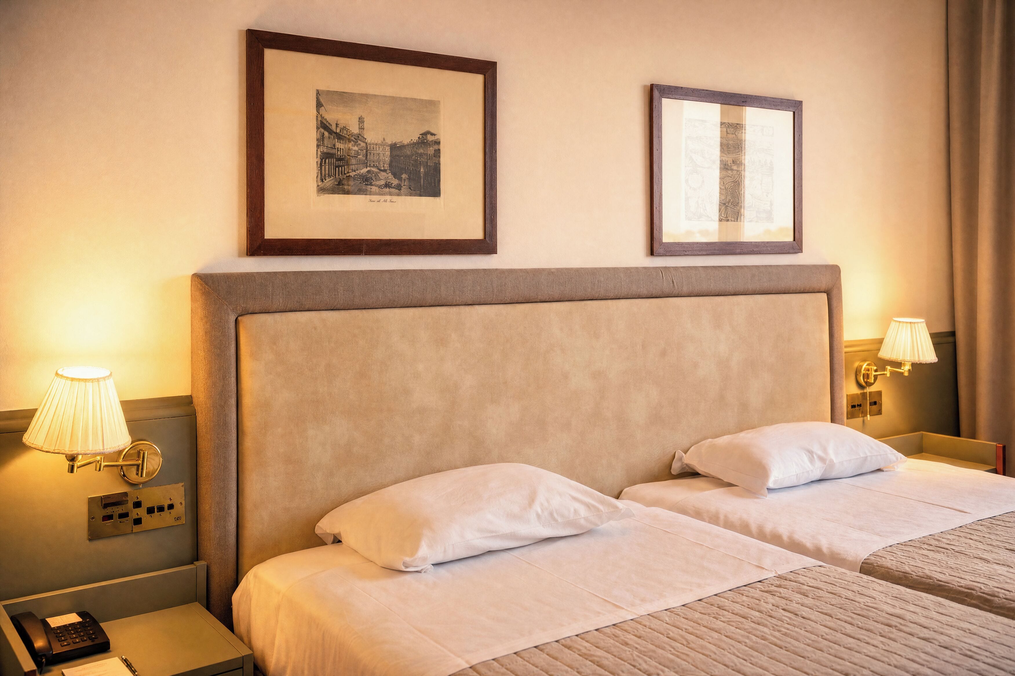 A hotel room with two beds, a nightstand with a phone, and two wall-mounted lamps.