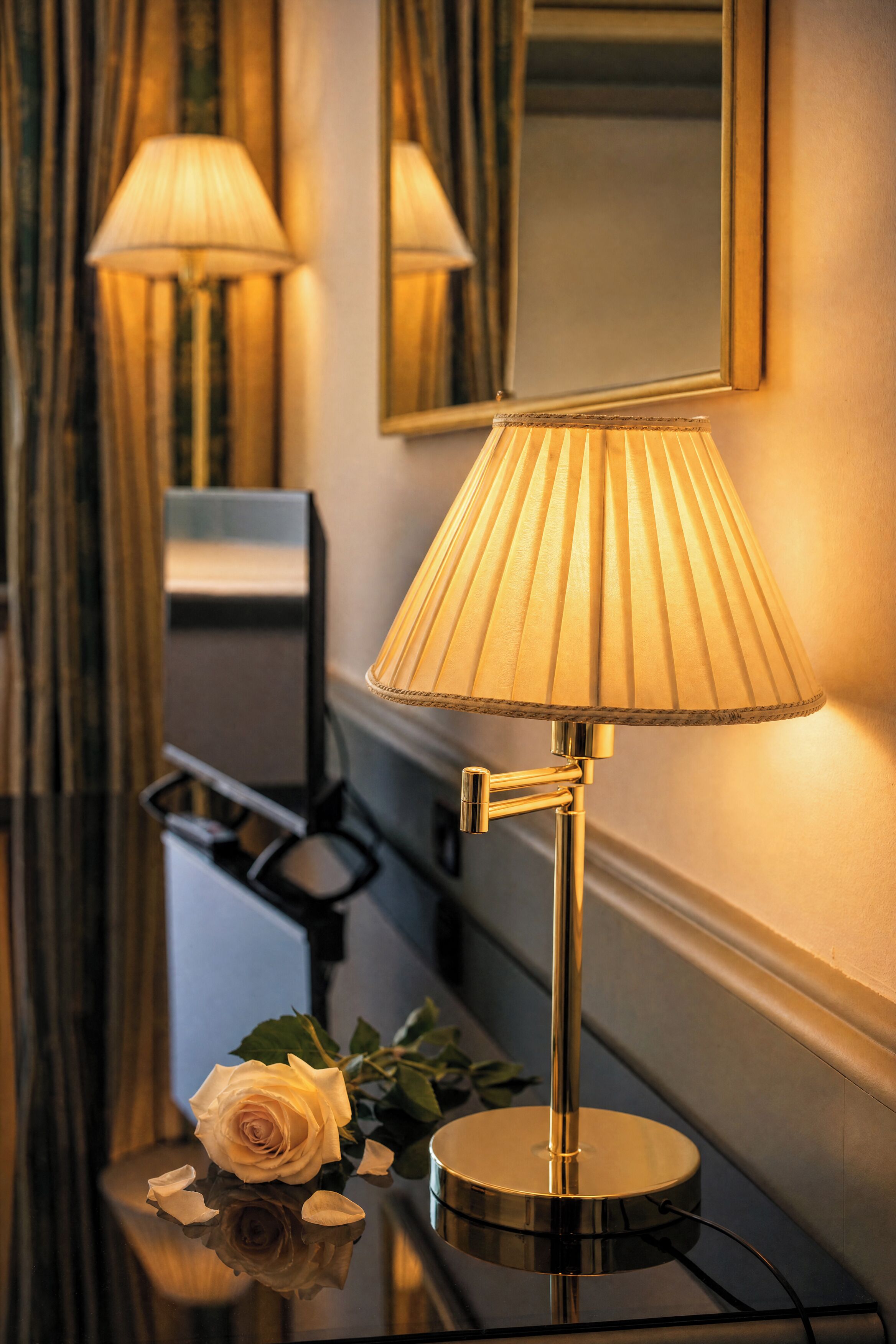 A lamp with a pleated shade on a reflective surface, a mirror on the wall, and a white rose on the table.