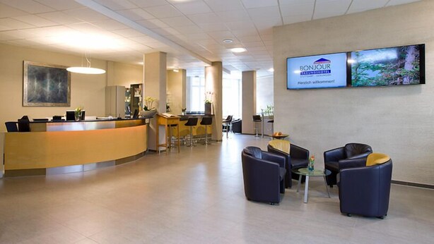 En hotellobby med en böjd receptionsdisk, en bardel och sittplatser med en tv-skärm som visar texten ”BONJOUR TAGUNGSHOTEL”.