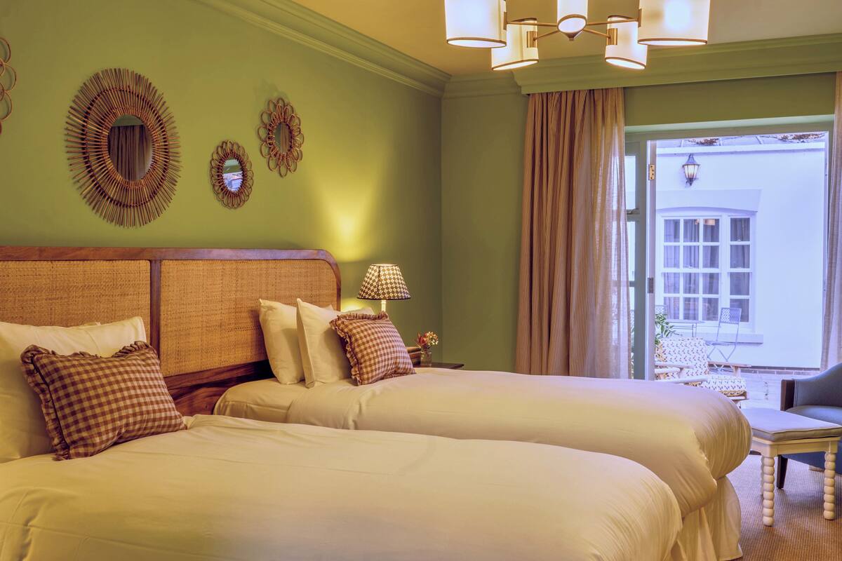 A hotel room with two beds, a nightstand with a lamp, and a view of the outside.