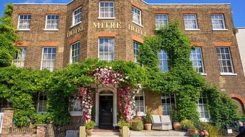 Een historisch bakstenen gebouw met de naam "MITRE HOTEL" duidelijk zichtbaar. Het heeft een klassieke entree met een decoratieve boog en weelderige beplanting.