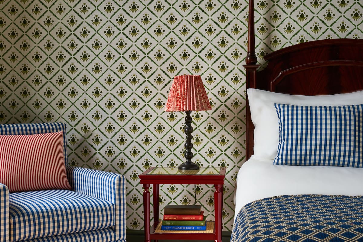 A neatly arranged bedroom with a patterned wallpaper, a wooden bed frame, a red nightstand with books, a blue and white checkered chair, and a red lamp.