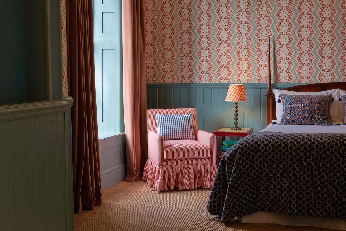 A bedroom with a bed, a chair, a lamp, and patterned wallpaper.