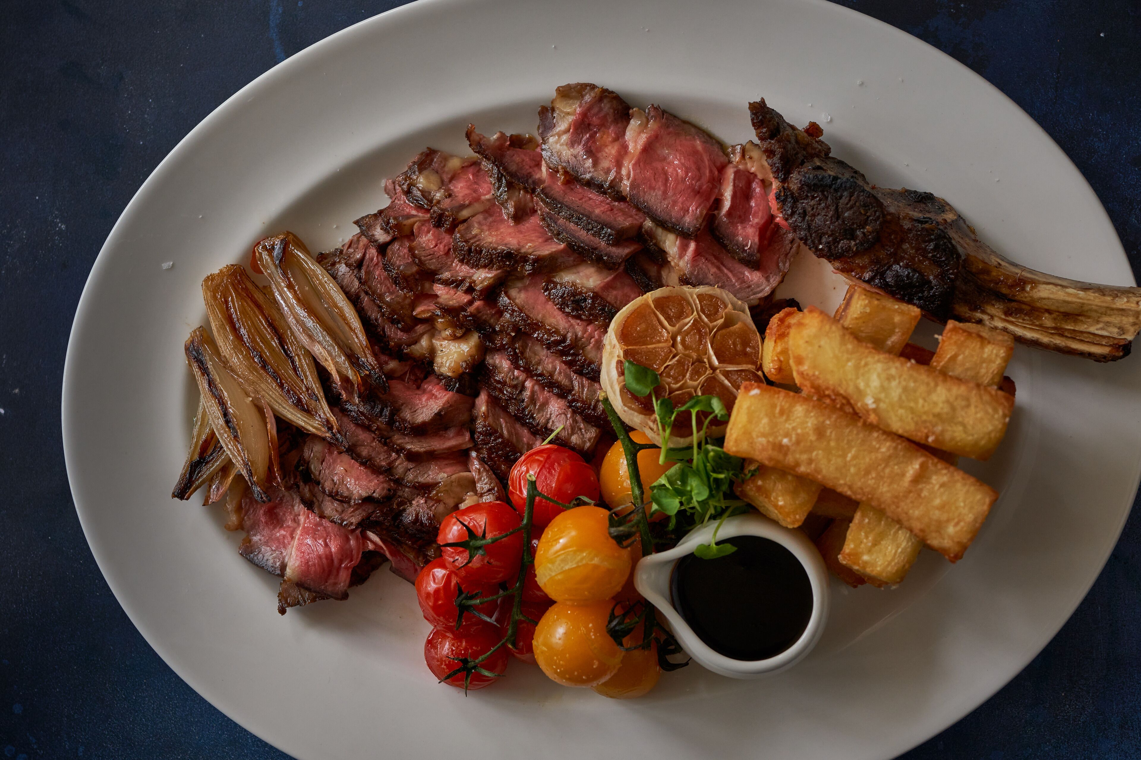 A plate of sliced steak, roasted potatoes, cherry tomatoes, and a small bowl of sauce.