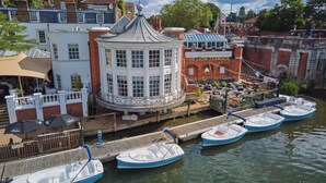 A waterfront hotel with a central tower, a restaurant terrace, and moored boats. - The Mitre, Hampton Court (East Molesey)