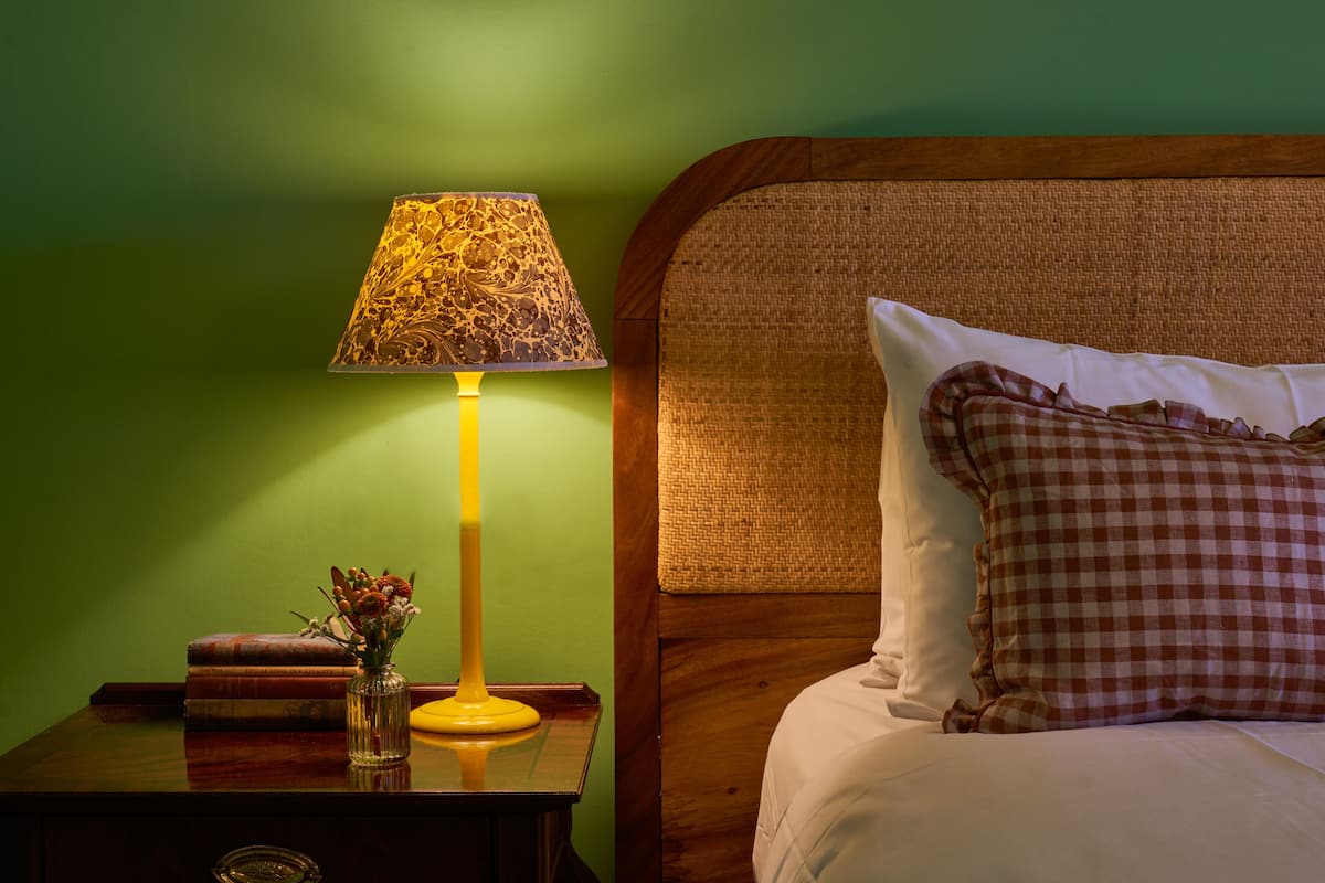 A neatly made bed with a checkered pillow and a wooden headboard. A bedside table with a lamp and a vase of flowers is visible.