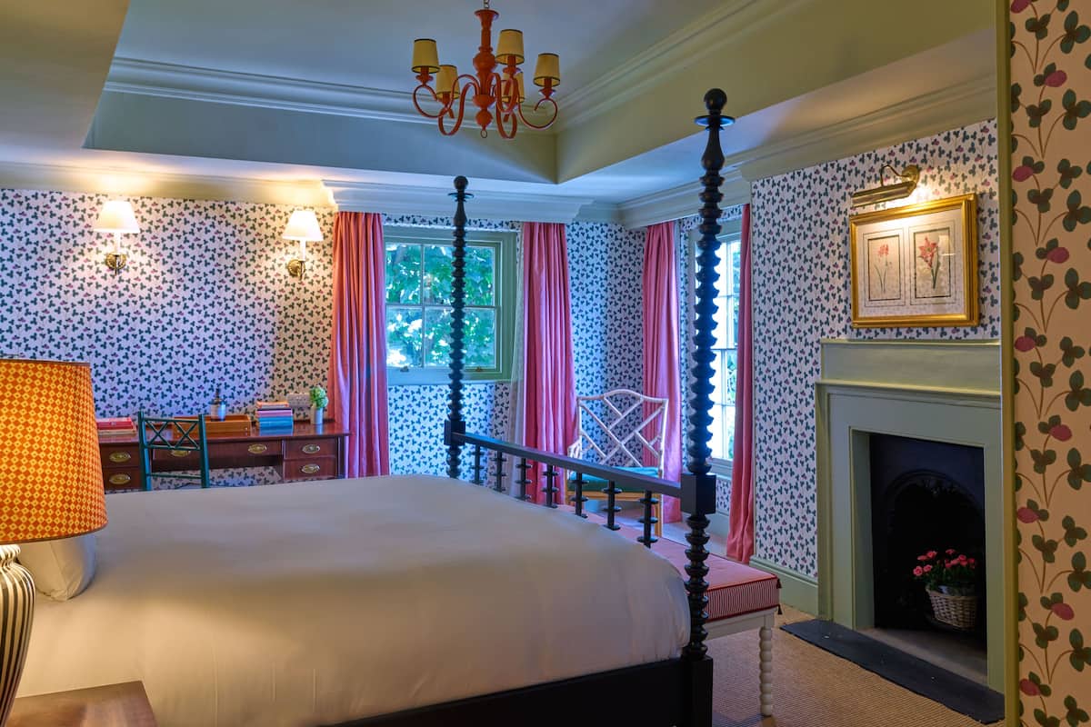 A four-poster bed with a white bedspread, a chandelier, a fireplace with a basket of flowers, and patterned wallpaper.