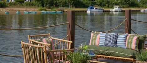 A wooden deck with a wicker sofa and chairs, overlooking a calm river with boats and houses in the background.