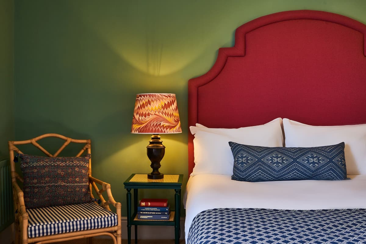 A neatly made bed with a patterned blue blanket, a red headboard, and a matching pillow. A wicker chair with a patterned cushion and a small table with a lamp and books are also visible.