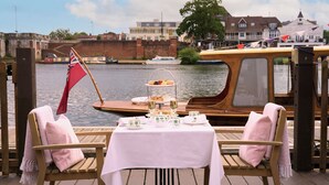 A wooden boat with a table set for tea service, overlooking a river with buildings in the background. - The Mitre, Hampton Court (East Molesey)