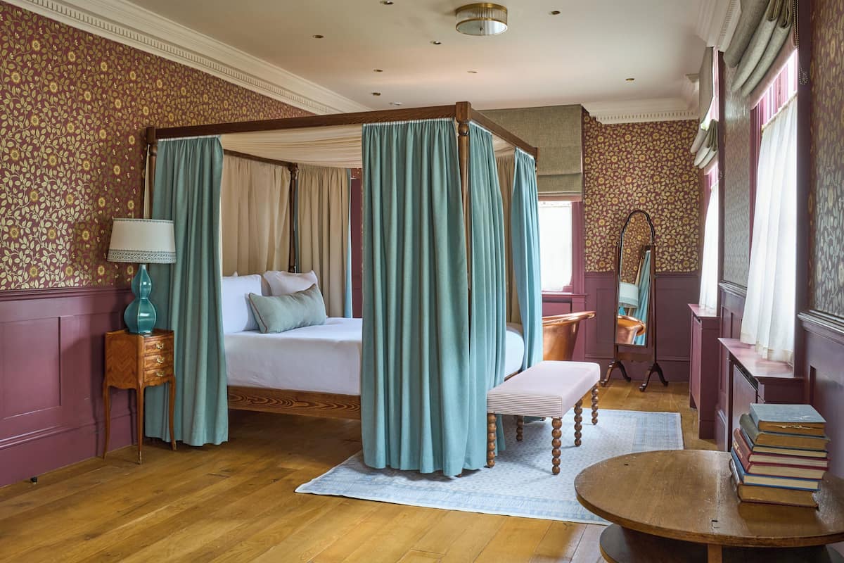 A four-poster bed with teal curtains, a bedside table with a lamp, and a stack of books on a round table.