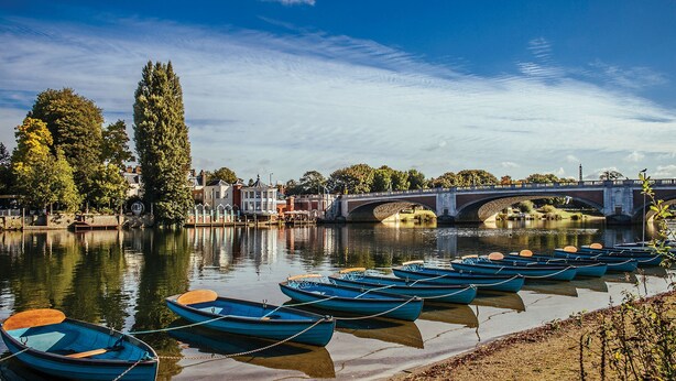 A river with boats docked along the shore, a bridge in the background, and buildings on the opposite bank. - The Mitre, Hampton Court (East Molesey)