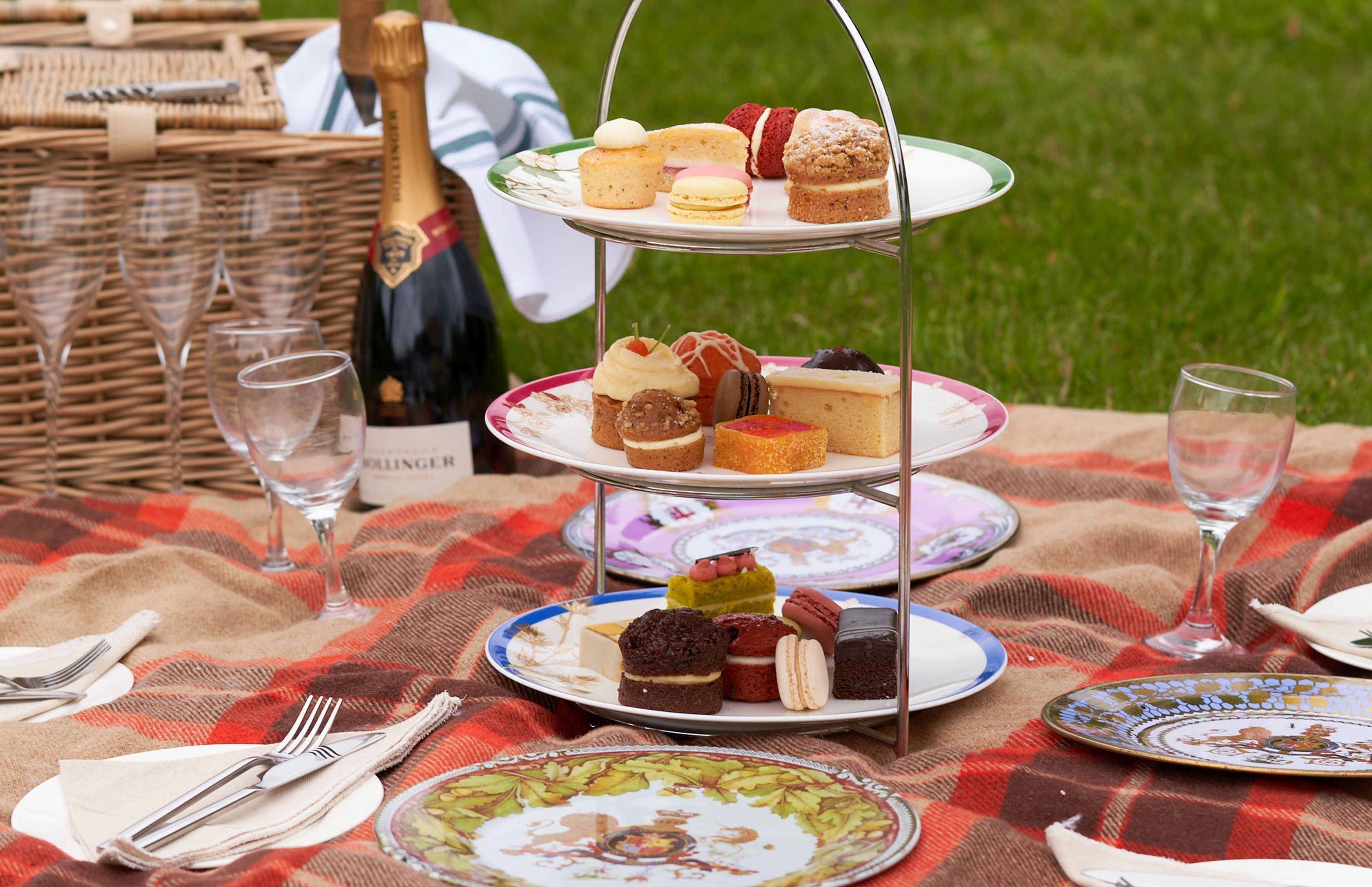 Een picknickopstelling met een etagère voor desserts, wijnglazen en een champagnefles op een geruit tafelkleed.