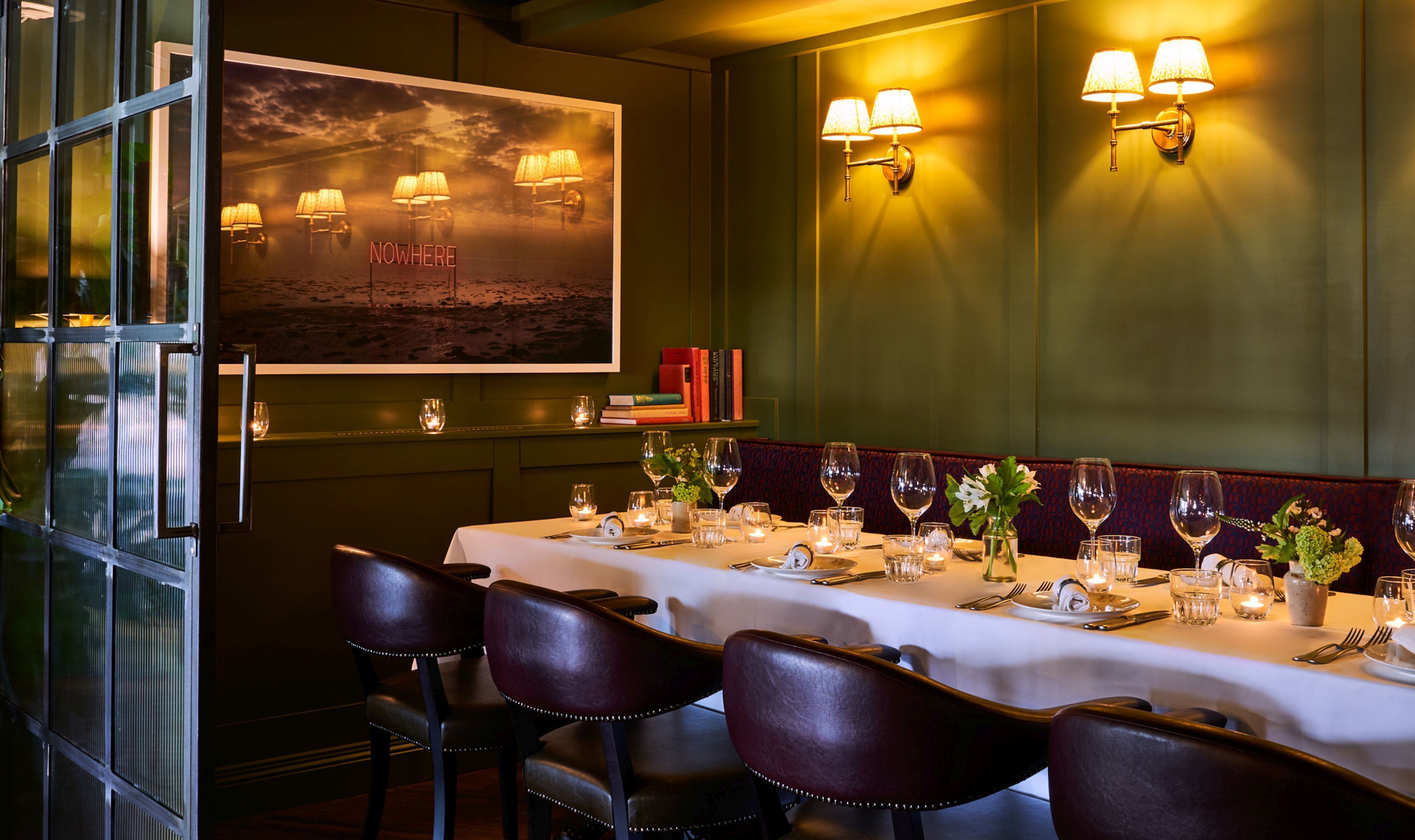 Un restaurant avec une longue table dressée pour le repas, un tableau accroché au mur et des lampes fixées au mur.