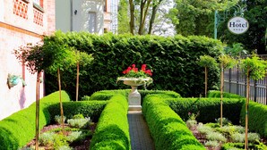 Um jardim bem cuidado, com um canteiro central de flores, um caminho delimitado por sebes e o letreiro de um hotel.