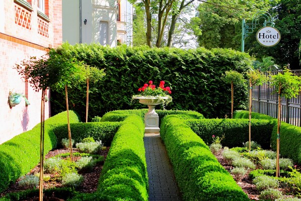 A well-manicured garden with a central flower bed, a hedge-lined pathway, and a hotel sign.