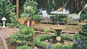 Um pátio com jardim, com mesa e cadeiras, cercado por diversos vasos de plantas e uma pequena fonte.