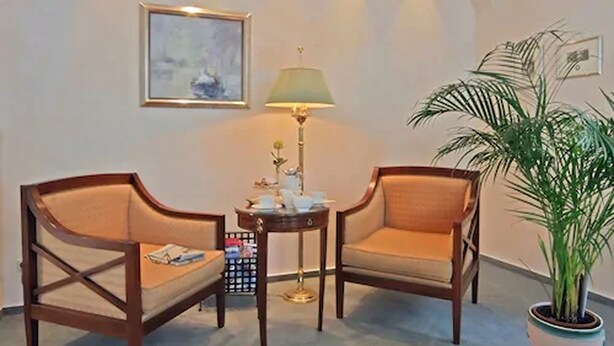 Una habitación con dos sillas de madera con cojines, una mesita redonda con una lámpara, un cuadro en la pared y una planta en maceta.