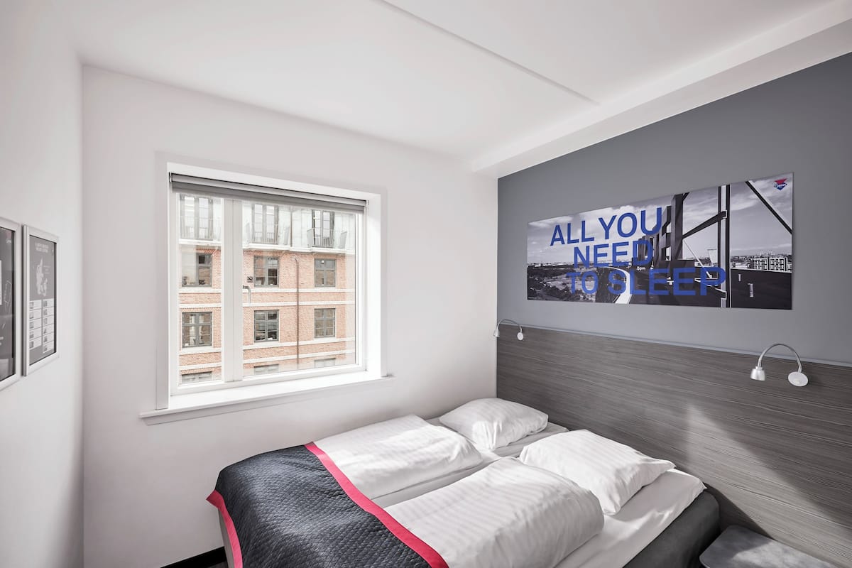 Une chambre d’hôtel moderne avec un grand lit, une fenêtre donnant sur un immeuble en briques et une œuvre d’art murale comportant du texte.