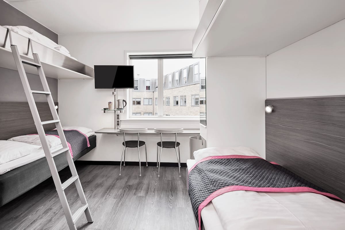 Une petite chambre avec des lits superposés, un bureau, une télévision et une fenêtre donnant sur un immeuble.