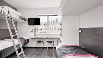 Een kleine kamer met stapelbedden, een bureau, een televisie en een raam met uitzicht op een gebouw.