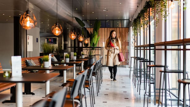 A modern cafe with wooden tables and chairs, hanging pendant lights, and potted plants.